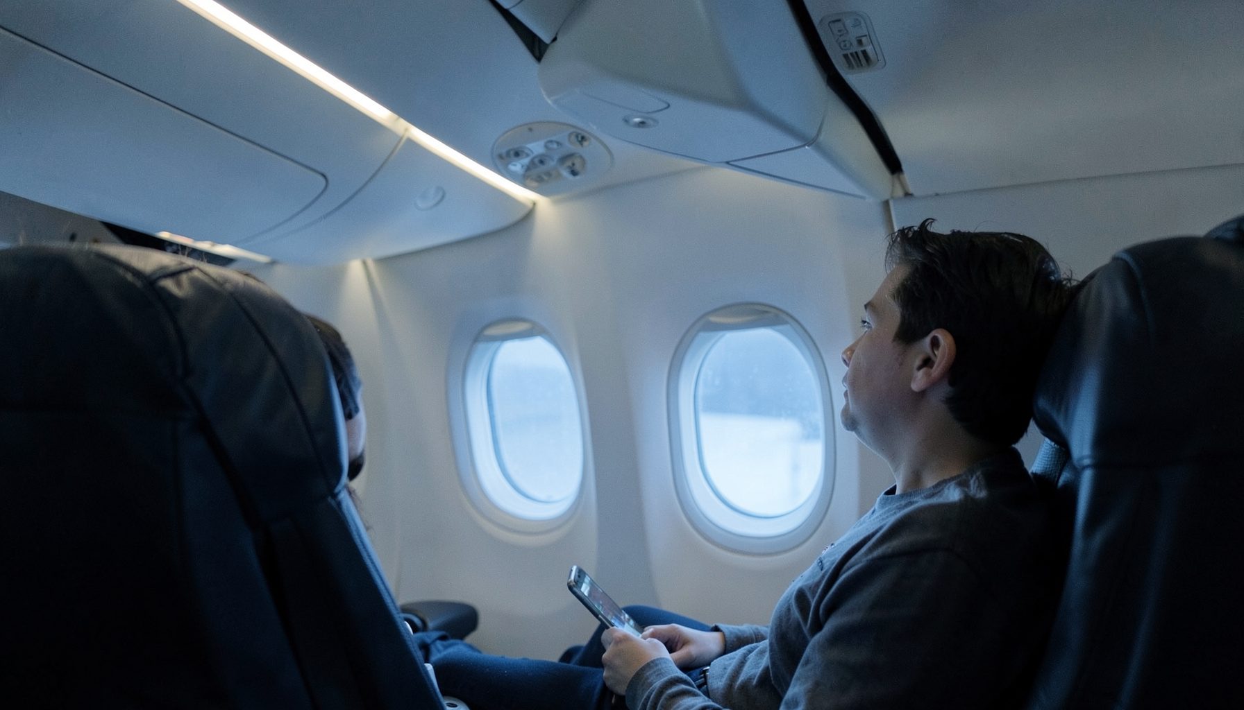 Airplane cabin interior with soft overhead lighting, a passenger looking out the window, calm blue-gray atmosphere