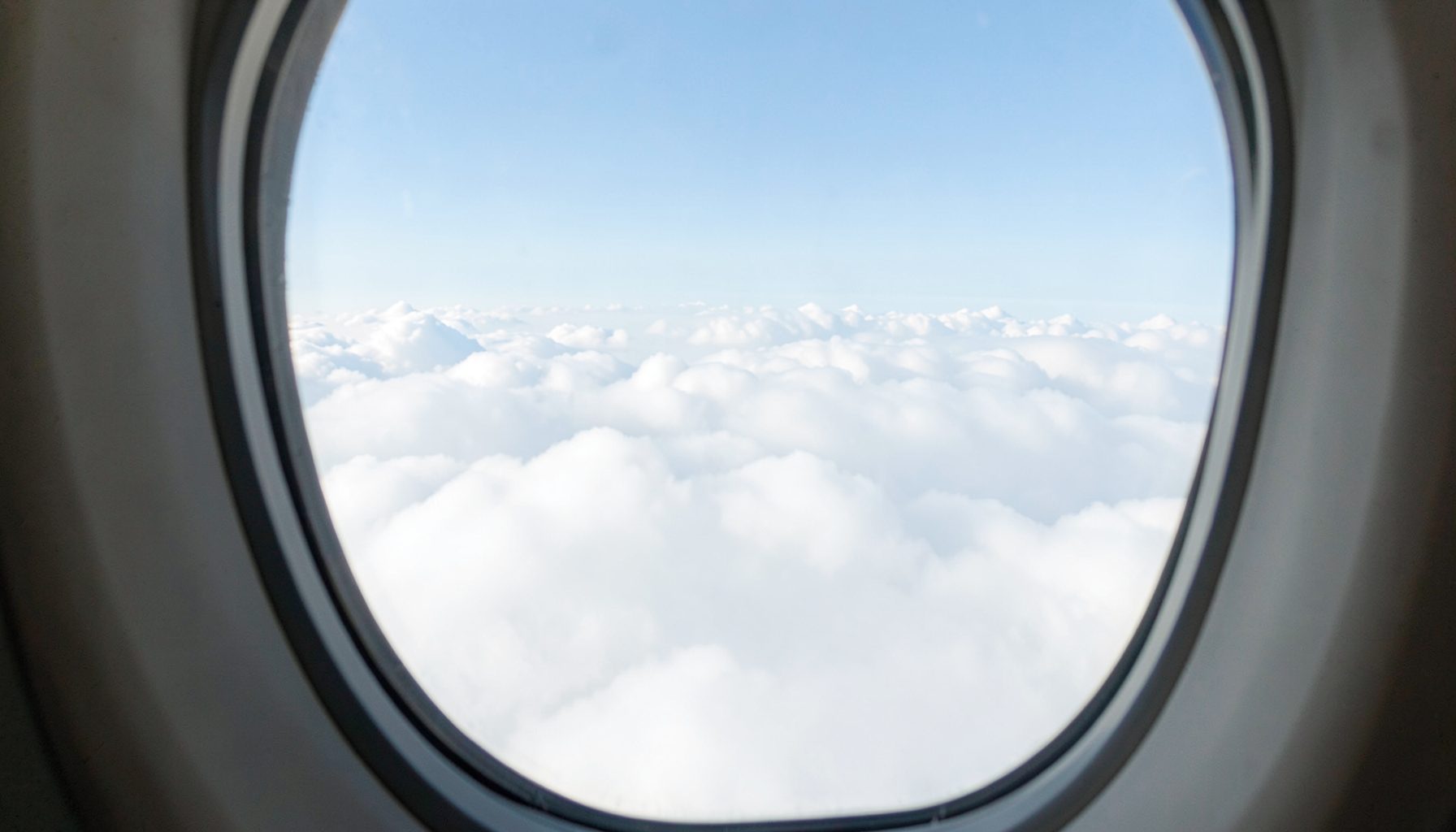 Airplane window view above soft white clouds against a pale blue sky, calm travel atmosphere