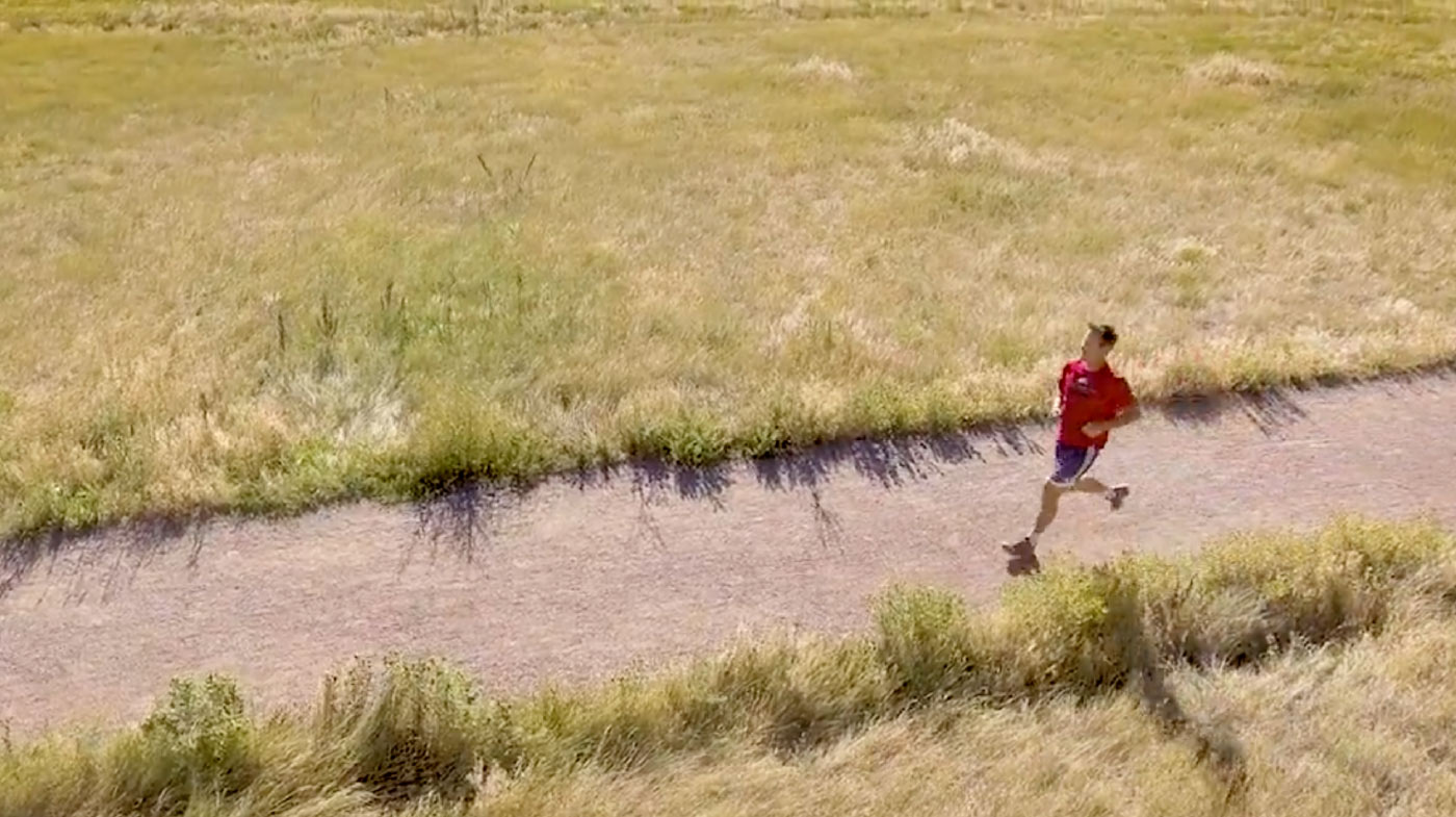 Aerial view of a person jogging in a red shirt along a dirt path through a grassy field, warm green and tan tones, active outdoor lifestyle